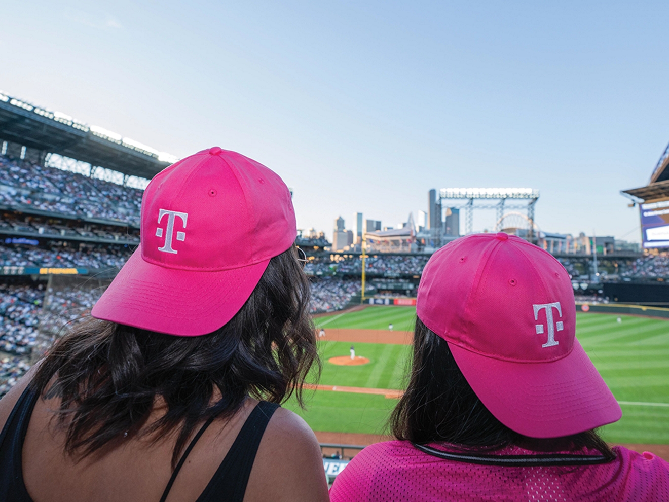 ​​Two friends watch a baseball game while wearing magenta T-Mobile baseball caps.​ 